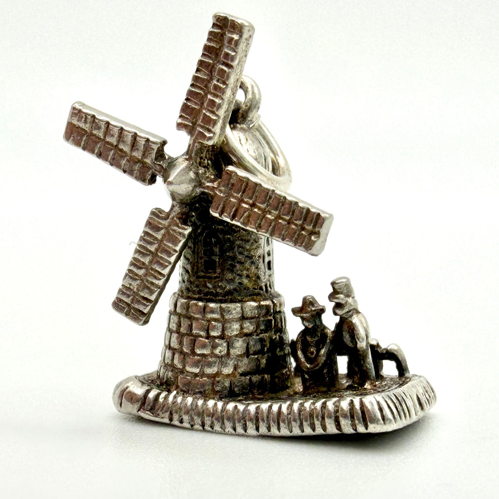 A silver-toned metal charm shaped like a windmill with two small figures and a dog beside it, photographed on a plain white background.