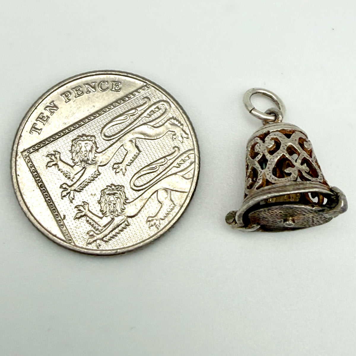 A silver filigree bell-shaped pendant charm shown next to a UK ten pence coin for scale, on a plain light gray background.