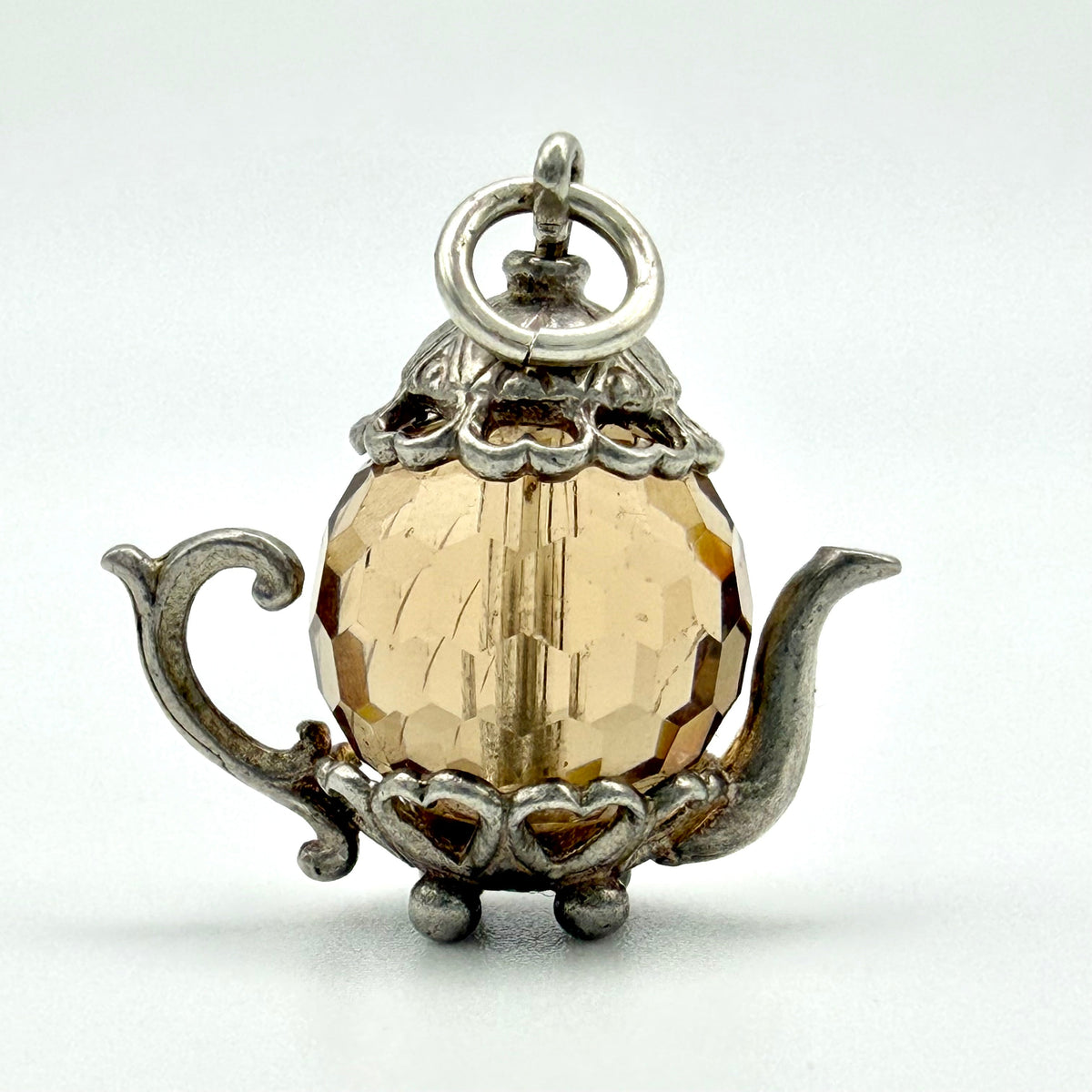 A vintage-style teapot pendant charm featuring a faceted amber glass sphere set in an ornate silver-toned metal frame, shown on a light gray background.