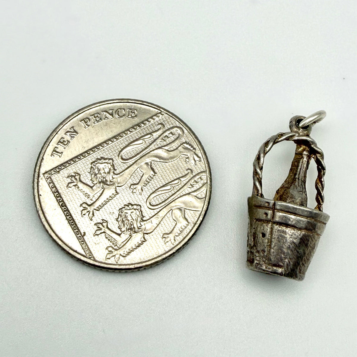 A small silver bottle-bucket pendant shown next to a UK ten pence coin for scale, on a plain light background.